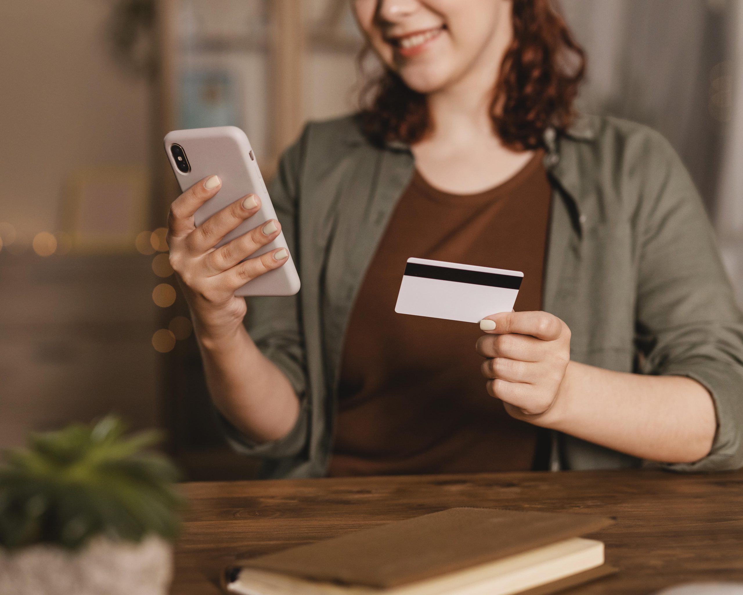 A smiling woman sits at a wooden table holding a smartphone in one hand and a credit cards in the other. She wears a brown shirt and a green jacket. A notebook and a small plant are on the table. The background is softly lit with warm, blurred lights, creating a cozy atmosphere.