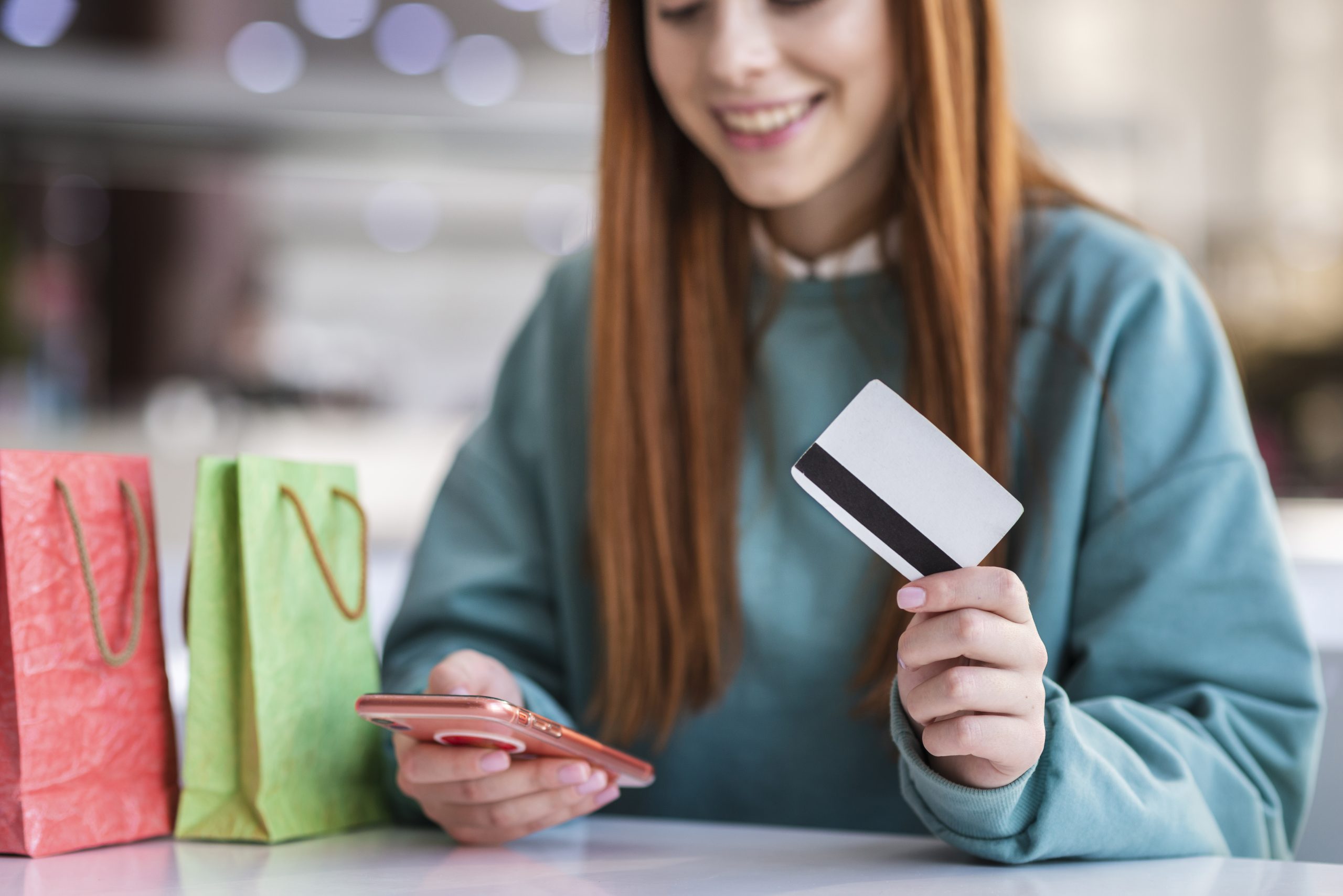 A person holding a debit card in one hand and using a smartphone in the other, surrounded by shopping bags, showcasing the convenience of using debit cards for online and in-store purchases.
