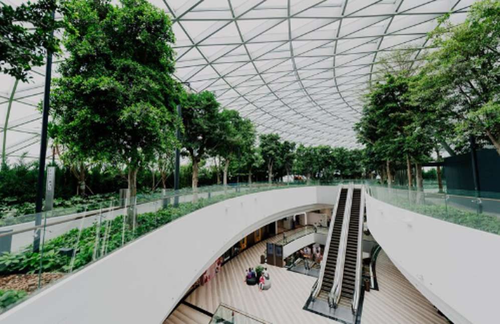 A spacious indoor public area showcasing green infrastructure, featuring abundant trees and plants integrated into the architecture, with natural light streaming through a glass-panelled roof and a central escalator connecting two levels.