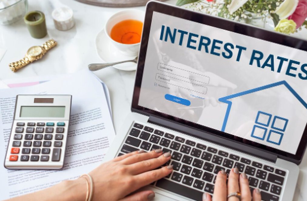 A person using a laptop displaying an online platform about interest rates, with a calculator, documents, and a cup of tea on the desk, symbolising financial planning and decision-making in the property market.